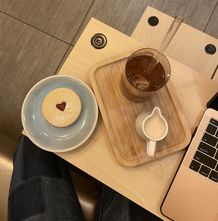 An iced coffee and a small container of milk on a tray beside a thumbprint cookie and a student's laptop.