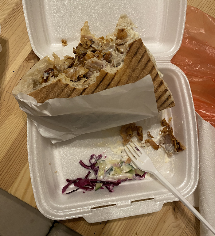 A takeout container of a grilled Gyro sandwich with a couple bites taken out of it. 