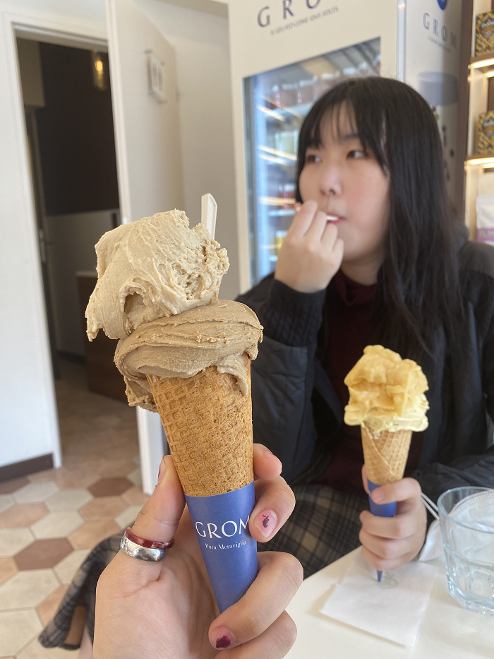 A student's hand holding a sugar cone with two scoops of coffee gelato. Another student eats their own gelato in the background. 
