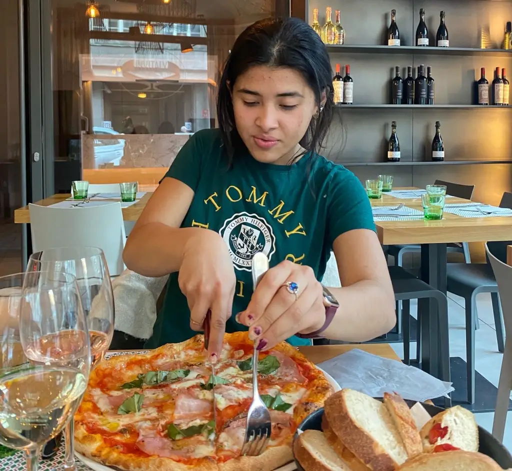 Gaby cutting into a pizza with a knife and fork in a restaurant.