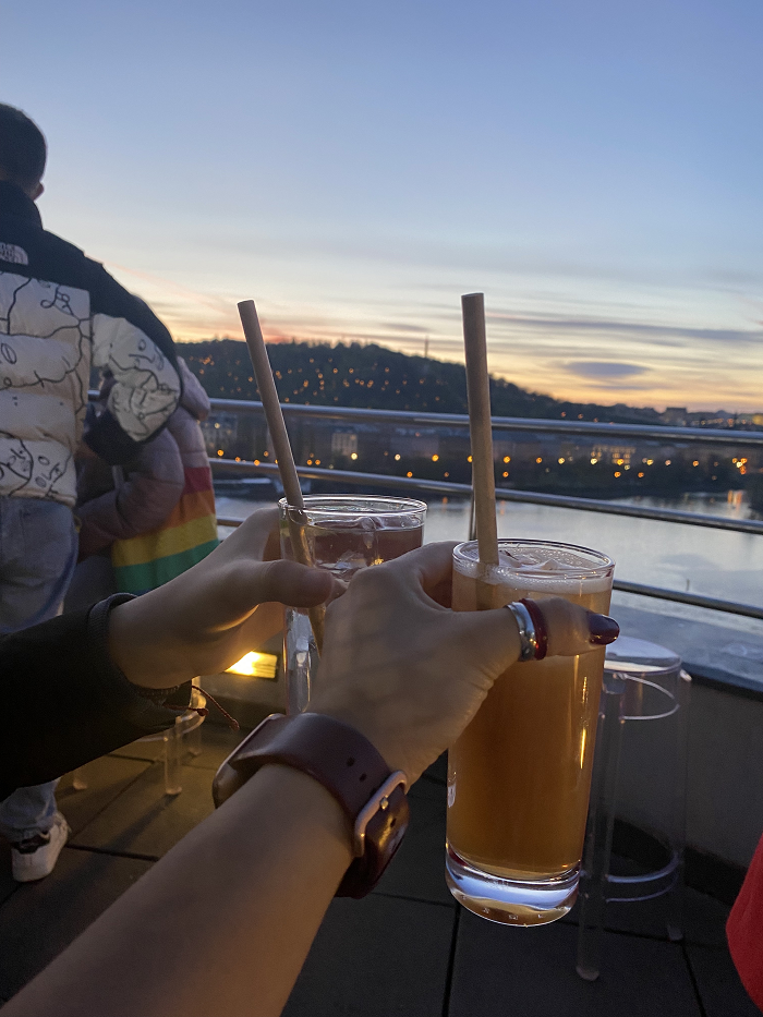 Two hands each holding a tall cocktail with a straw against the backdrop of a river and a hill at sunset. 