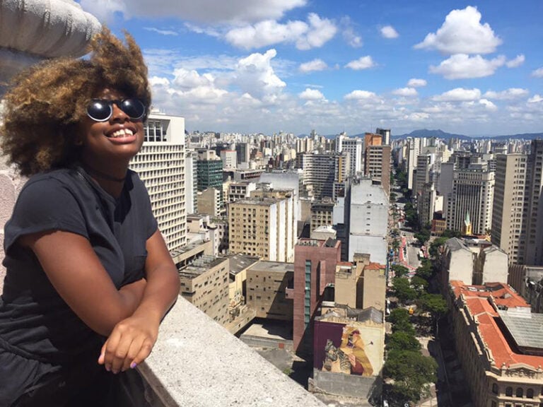 Student in Brazil stands on a balcony looking up at the sun, smiling, with the Sao Paulo skyline behind her