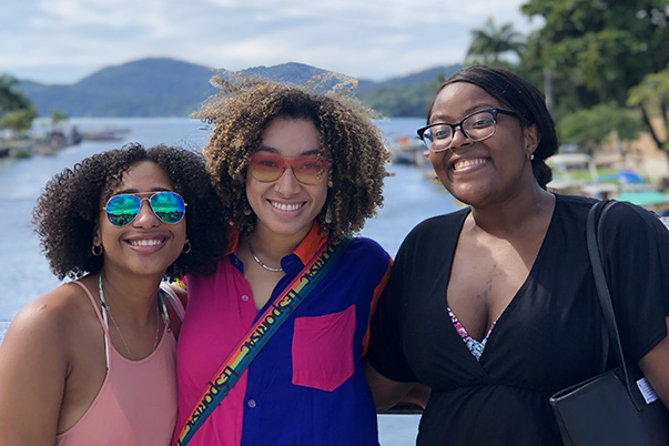 Three female students smile in front of a beach town in Brazil