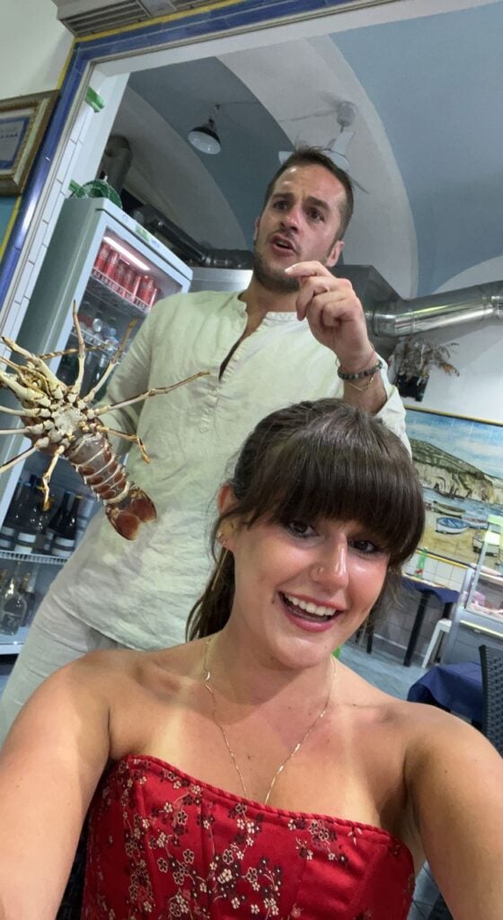 Grace taking a selfie with a man holding a lobster in the background 