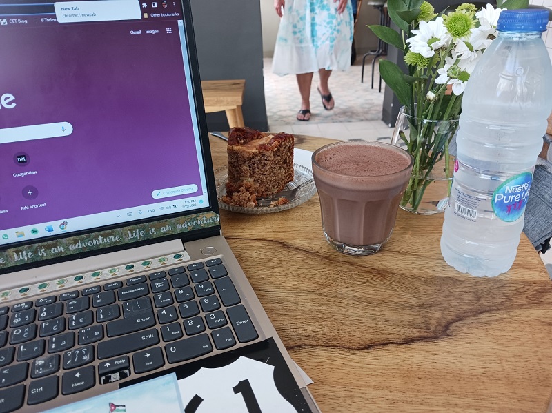 A laptop next to a slice of cake, a glass of coffee, and a water bottle at a café table 