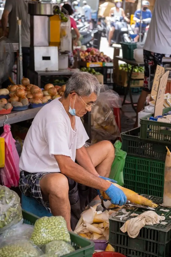 A local market vendor chops bamboo shoots with a cleaver atop plastic produce baskets.