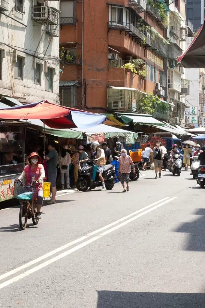 A bustling neighborhood wet market as seen from across the street. People on foot and motorcycles are passing in the foreground.  