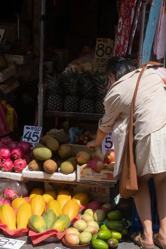Elissa's roommate is bending over to select mango from the colorful produce stand. 