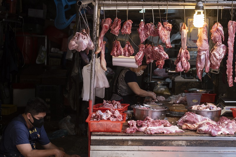 A local market butcher. Different cuts of meat hang from the top railing and more sit on the table below. Two masked vendors are nearby. 