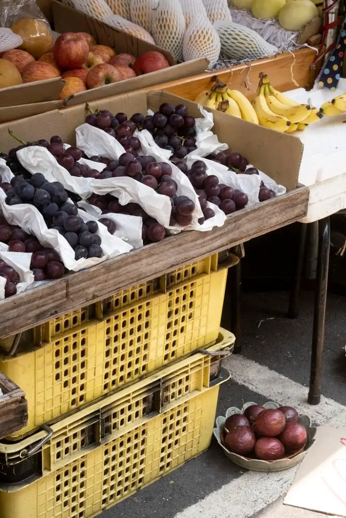 Produce on display at the market above yellow produce baskets. There are grapes, bananas, and pomegranates. 