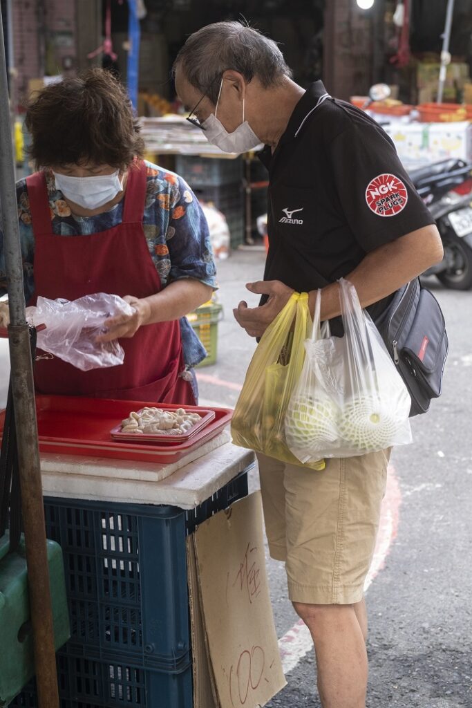 A masked man holding plastic bags of produce watching a masked vendor package his produce. 