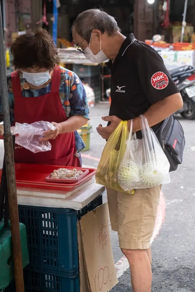 A masked man holding plastic bags of produce watching a masked vendor package his produce. 