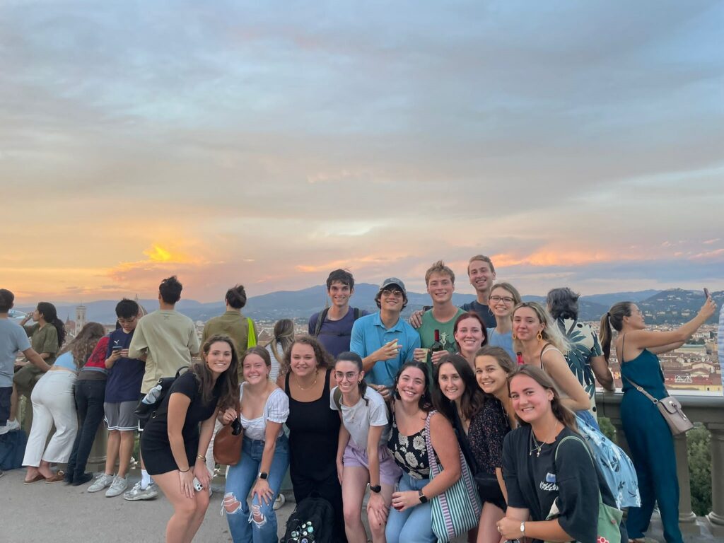 A group of students posing for a photo in a highly trafficked tourist spot during sunset. Other tourists are seen admiring the scenery and taking photos in the background. 