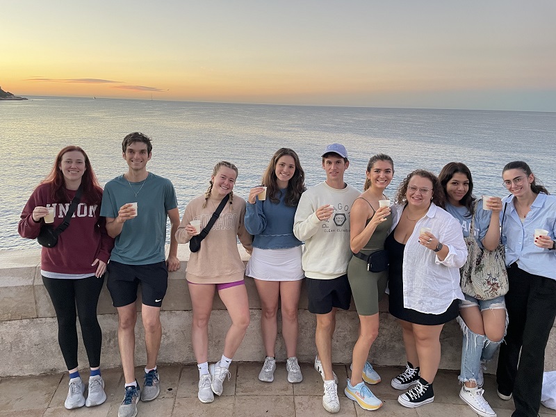 Nine students holding small white cups and smiling while posed against a half wall before the ocean. 