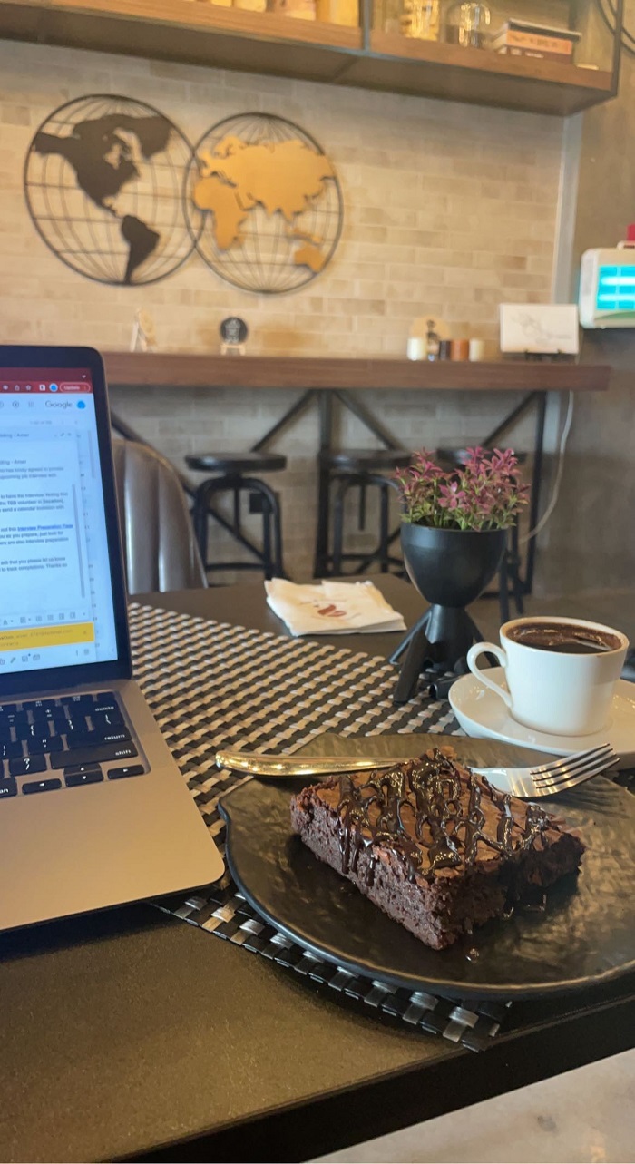 A slice of a cake atop a café table beside a coffee and an open laptop.