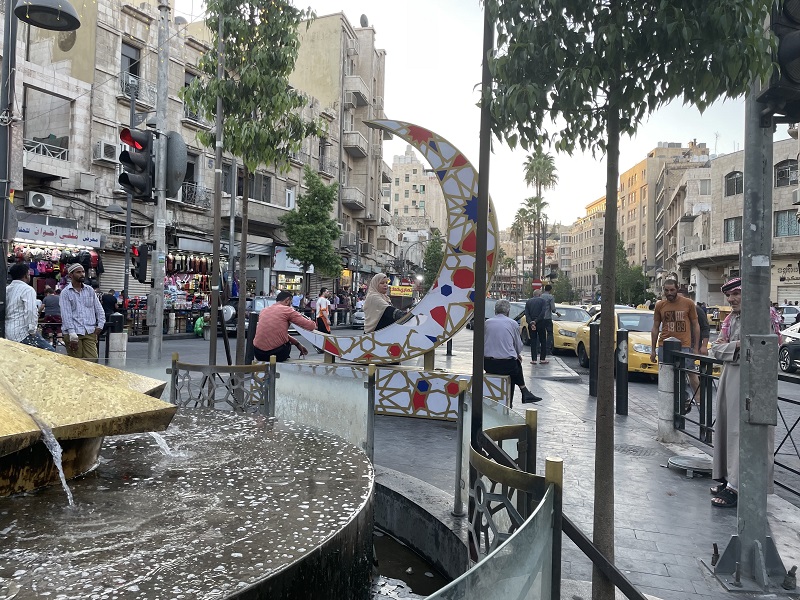 A small plaza in Amman. People are seated casually, some are standing nearby crossing the street. Shops can be seen in the background. 