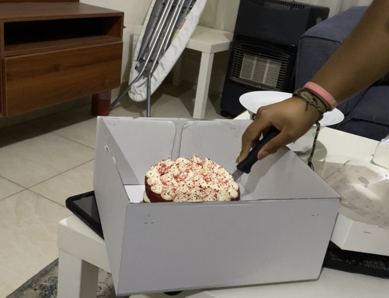 A student's hand cutting into a small cake in a box atop a table.