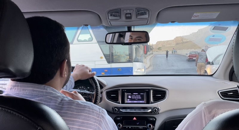 The view from the backseat of an Uber. A male driver is seen driving the vehicle on the left side, the car's interior is mostly gray.