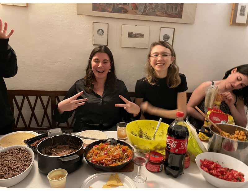 Three students at a dining table with a taco station including peppers, beef, and tomatoes