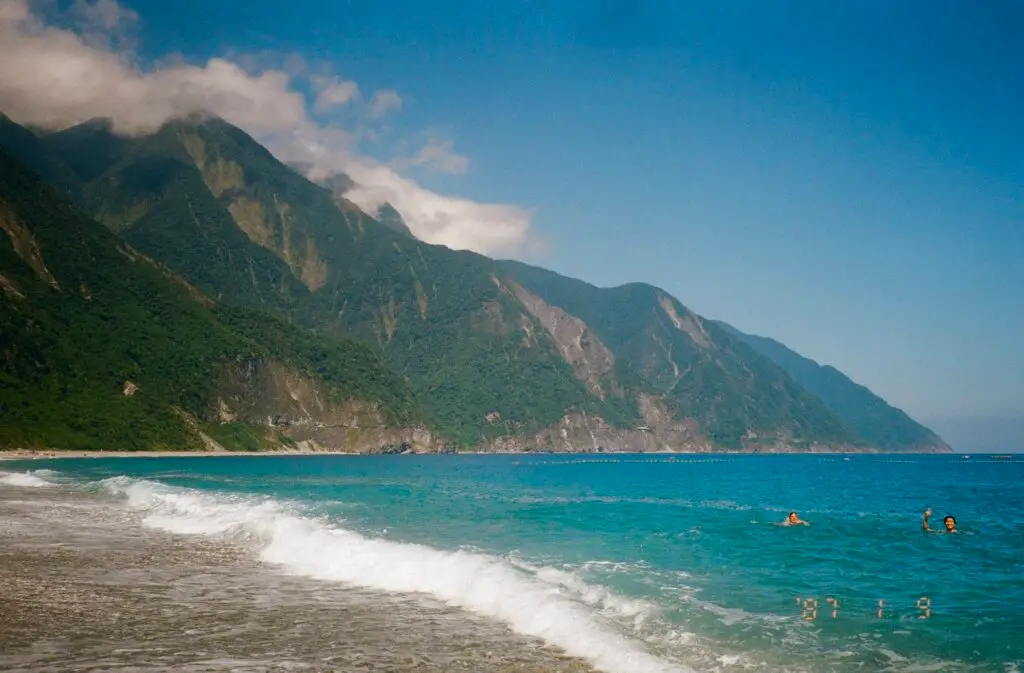 A wise shot of turquoise waters breaking into white sea foam along a gray sand beach. The background has steep mountains on a partly cloudy day.