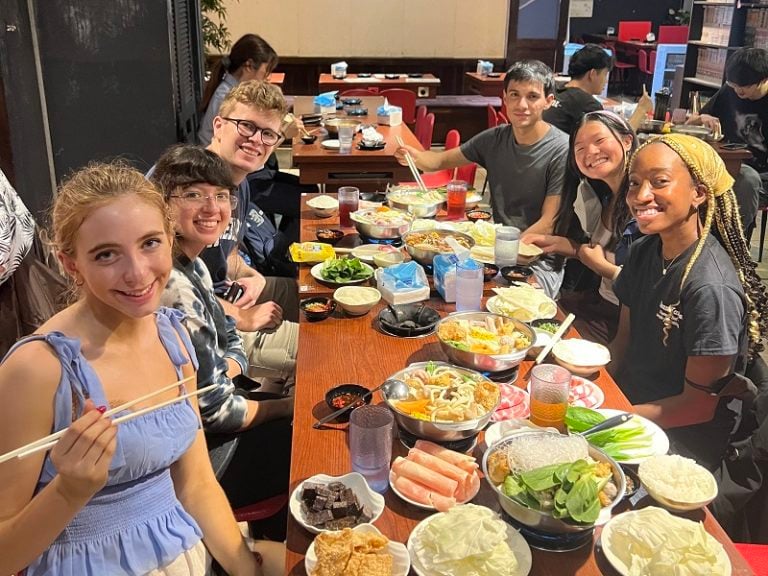 A group of students around a long table at a hot pot restaurant posing for a photo. The table has many small plates of raw foods.