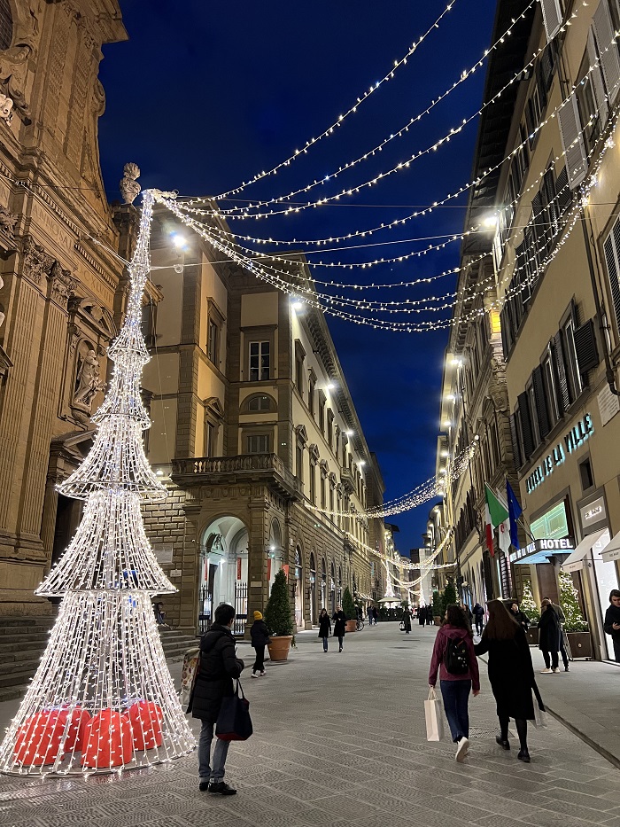 A street in Florence during the Christmas holiday season with white lights, shoppers, and white Christmas tree