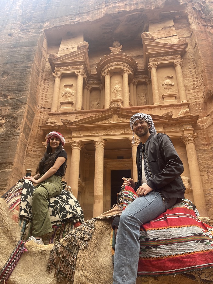Two students smiling while riding on the backs of camels as they pass the Al-Khazneh Treasury 