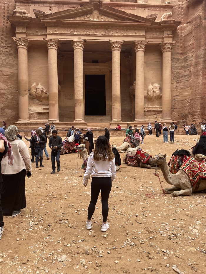 A student, locals, and others standing in front of Petra with camels sitting nearby.
