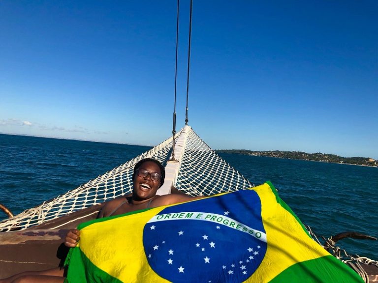 A student smiling and holding the Brazilian flag while on a boat during a sunny day.