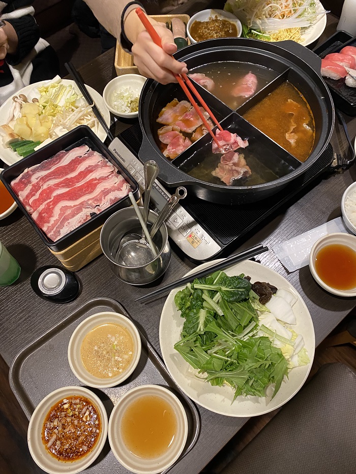 A hand holding red chopsticks, grabbing pieces of meat. The table is covered with hot pot items including vegetables, meats, sauces, and more.