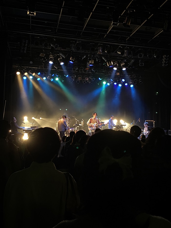 Roth Bart Baron performing at a live house show in Osaka, Japan in front of a crowd at an indoor venue. Multiple band players on the stage with green, yellow, and blue lights.