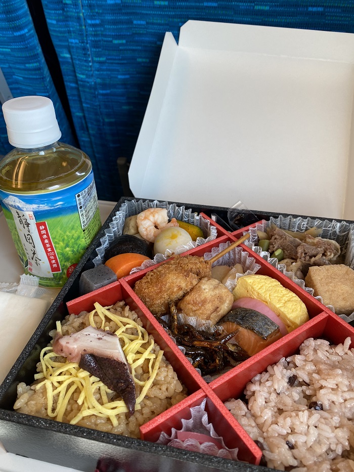 A bottled drink to the left of a bento box filled with rice, seafood meat, eggs, and more on a train ride tray