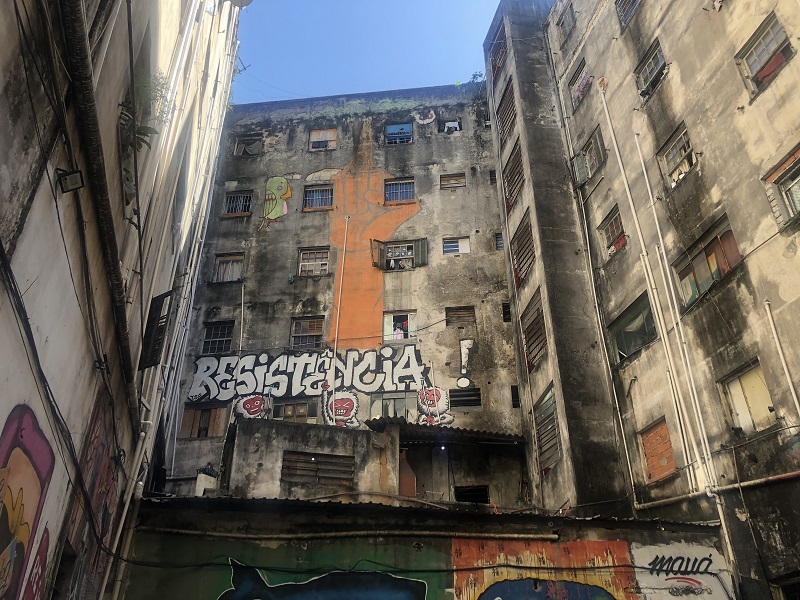 A run-down building called Ocupa Mauá in Brazil with wall art, such as the word RESISTENCIA! written in white and an orange clenched fist symbol of resistance