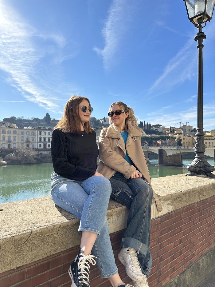 Two CET Florence students with sunglasses sitting along  the ledge in front of Arno River on a sunny day