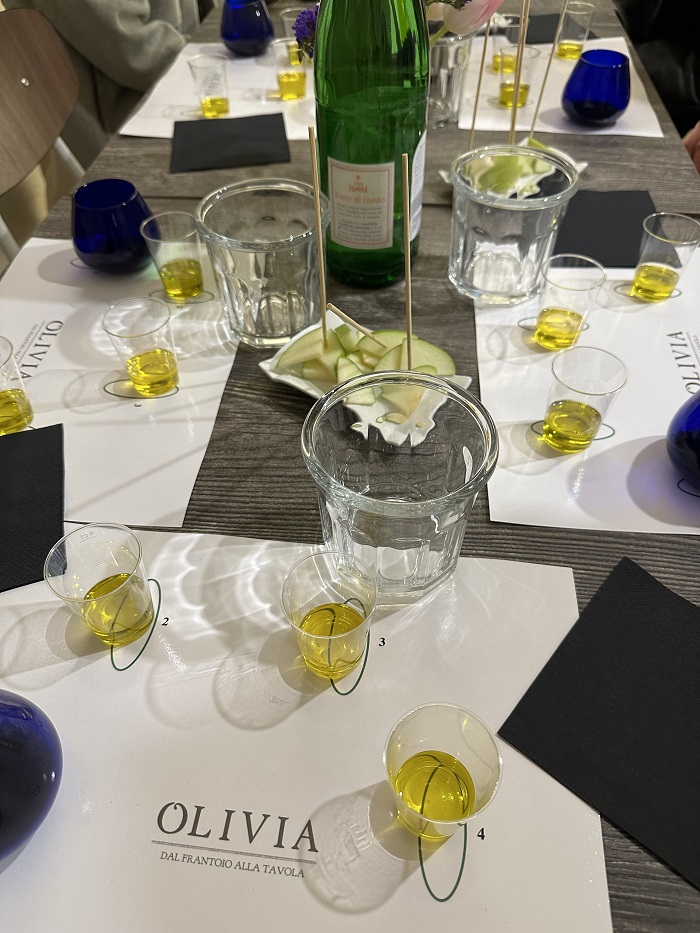 Little cups filled with yellow oil numbered on paper placemats, thinly green apple slices in the middle, and black napkins on a table