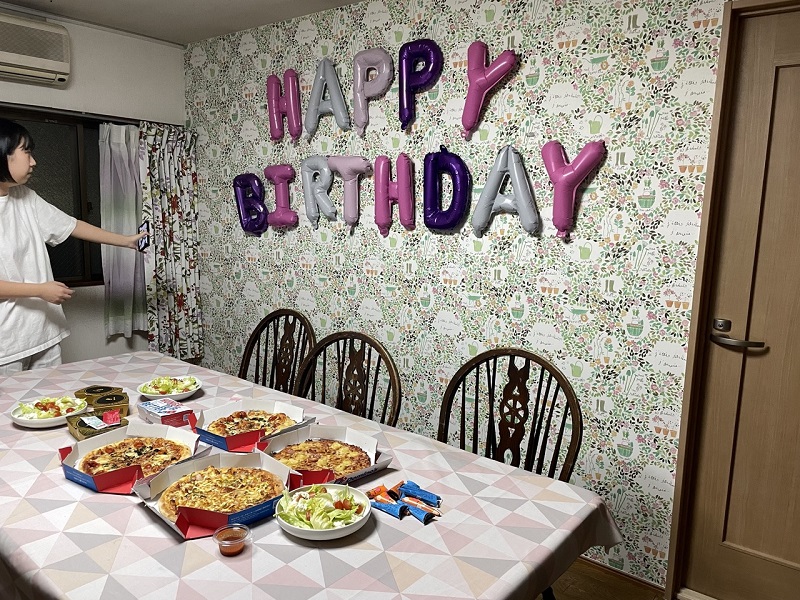 A Japanese housemate in a room setting up for a birthday party with pink, grey, and purple letter balloons on the wall and pizzas on the table.