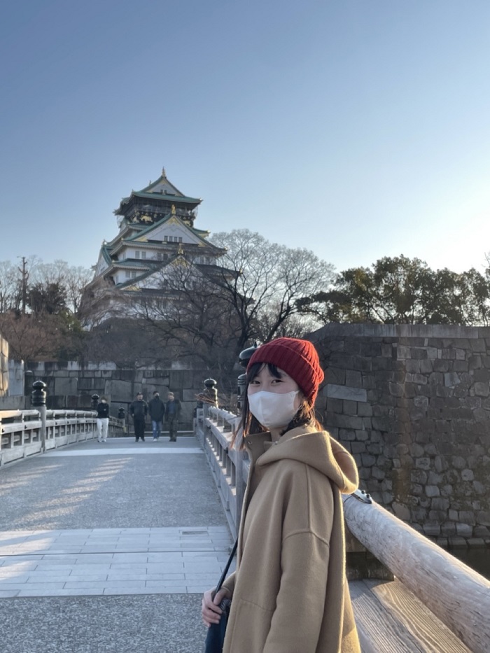 A CET Japan student outside wearing a red beanie and tan shirt, standing in front in Osaka Castle Park
