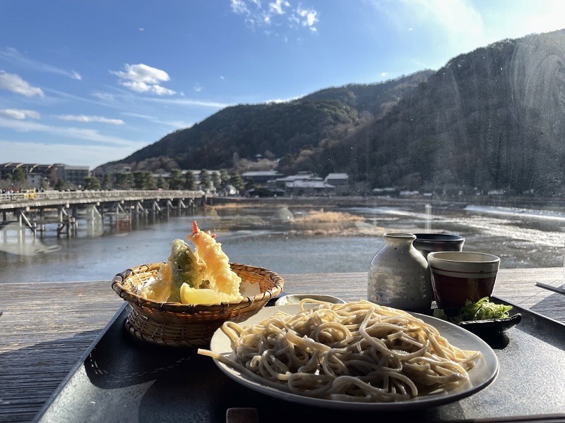 A bowl of shrimp tempura to the left, a plate of soba noodles in the middle, and tea to the right, all in front of a view of Arashiyama in Kyoto