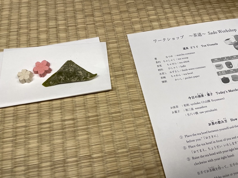 Tea snacks shaped in pink and white flowers on a napkin to the left and a piece of paper with information about the tea ceremony workshop in Japanese and English.