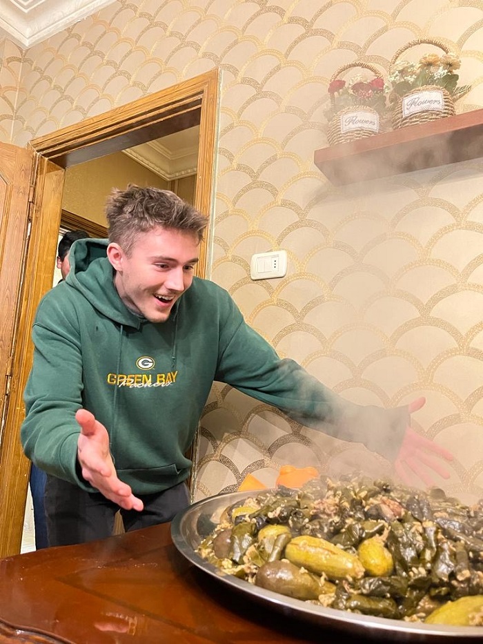 A CET Jordan student smiling excitedly with opened arms at a large dish of steaming dolma, Jordanian stuffed grape leaves food, on a table
