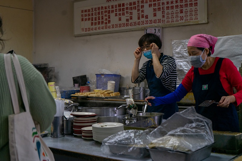 Two women in an open kitchen wearing masks in Taiwan. One woman is fixing her glasses while the woman to the right holds a ladle in a pot. There are plates and large grey containers in plastics bags.