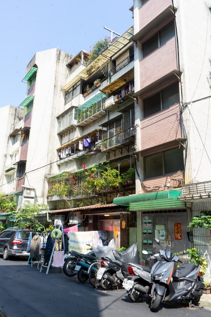 The side of an apartment complex in Taiwan with clothes and green plants hanging from patios. On the street there are more clothes hung up to dry, a parked car, and motorcycles.