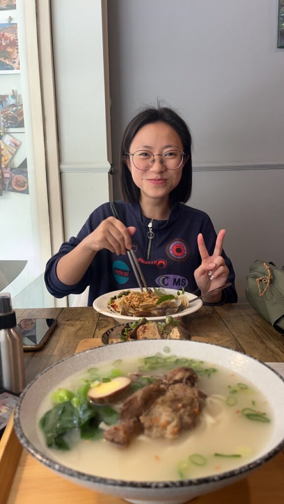 A CET Taiwanese housemate across the table, holding up a peace sign and chopsticks in her bowl. Closer to the camera, is a bowl of soup.