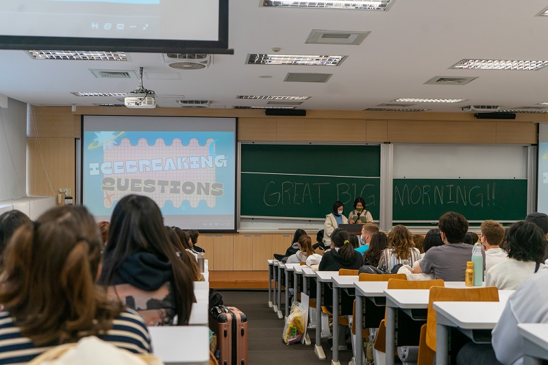 Many students sitting at desks in a classroom facing the front of the room towards two people standing behind a podium. The chalkboard has writing that says, "Great Big Morning!!" and to the left of the chalkboard is a screen with the words "Icebreaking questions."