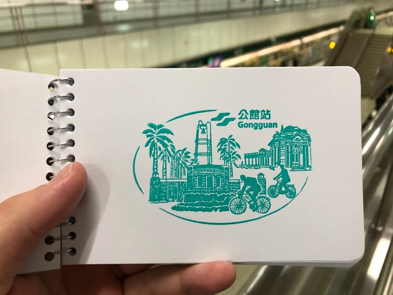 A green stamp of Gongguan station. The right-hand side of the stamp is the façade of the Musuem of Drinking Water and cyclists on the Gongguan Bikeway. The left-hand side has the entrance gate of the Royal Palm Boulevard and the Fu Bell.
