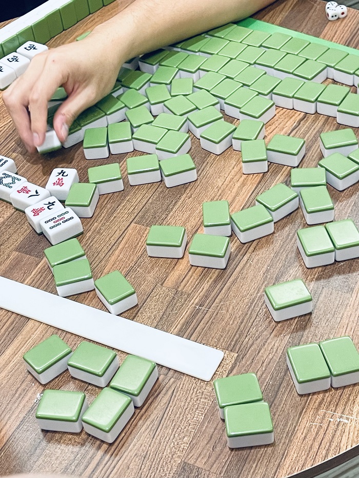 Green and white game pieces for mahjong scattered on a wooden table with some white game pieces and a hand picking up one of the pieces