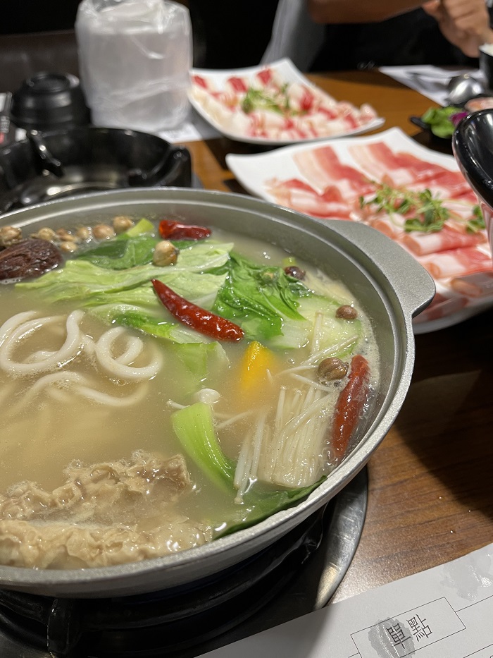 Light yellow hot pot soup with vegetables, noodles, and peppers with thinly sliced meat on white plates