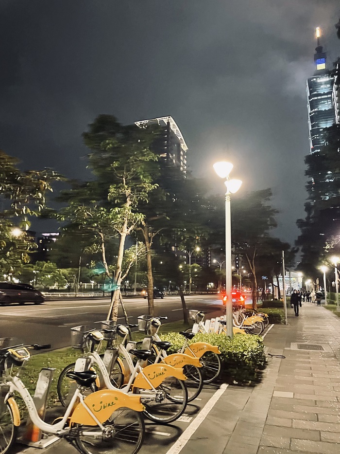Street in Taiwan at nighttime by Taipei 101 Tower with yellow bikes, driving cars, and people walking on sidewalks with lit up streetlamps