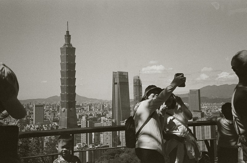 Black and white film photo of people along the railing of Xiangshan and a view of the Taipei 101 Tower and city landscape in the distance