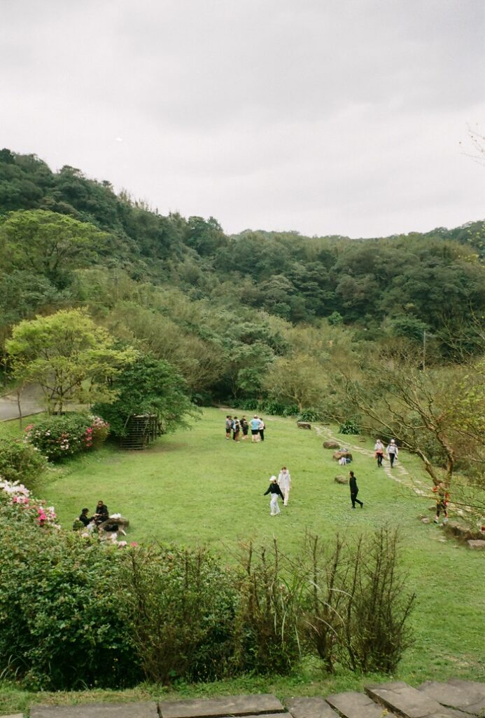 A view at Caoling Historic Trail in Taiwan with many trees surrounding the area and a patch of grass in the middle with people walking and sitting on it